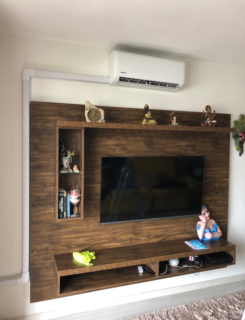 painel de marcenaria em madeira escura com TV, ar-condicionado, prateleiras com livros, biblets e pequenas esc