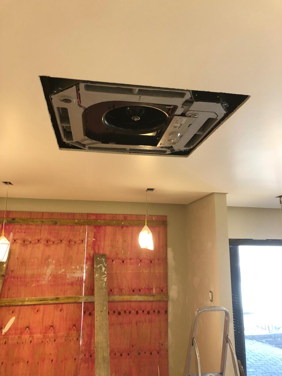 Equipamento de ar-condicionado tipo cassete aberto no teto, com ventilador visível, lâmpadas pendentes, parede